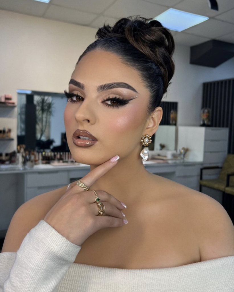 This look exudes sophistication with its meticulously applied makeup and classy updo. The eyes are defined by a bold cat-eye liner and enhanced with shimmering neutral eyeshadows. Cheeks are sculpted with contour and a touch of highlighter, while the lips shine in a glossy brown. The overall appearance is elegant and perfectly suited for formal gatherings.